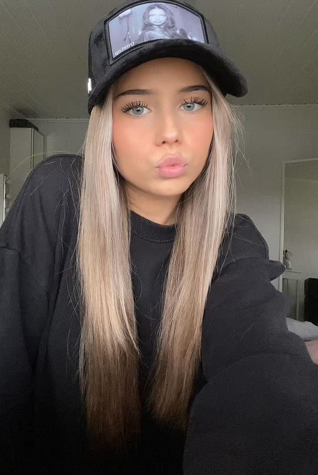 Woman with long blonde hair wearing a black hat with a printed image, posing indoors.
