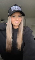 Woman with long blond hair wearing a black cap and black sweater smiles while posing indoors